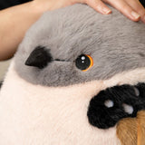 Close-up of a plush bird toy being held by a person's hand, showing gray and white fur, a black beak, a black-and-white spotted wing, and a yellow-rimmed eye.
