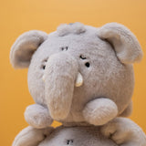 A close-up of a soft plush elephant toy in light gray, shown against a solid mustard-yellow background.