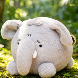 A round, soft plush elephant toy in light gray sits on green grass outdoors with a blurred forest background.