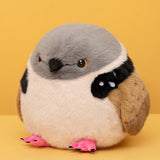 A round, fluffy bird plush toy with a gray head, cream belly, black wing patches, tan wings, and bright pink feet, photographed against a solid yellow background.