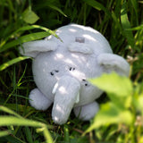 A white plush elephant toy lying on green grass outdoors, with sunlight and shadows across its fur.