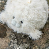 A fluffy white plush toy with a soft, shaggy texture, small dark bead eyes, and a pale gray stitched snout, resting on a rough stone surface.