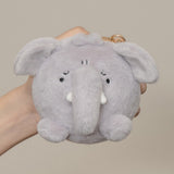 A small plush elephant keychain or pouch held in a hand against a plain warm beige background. The item is light gray with embroidered black eyes and nose, white tusks, and a small gold key ring at the top.