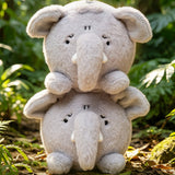 Two stacked plush elephant toys in light gray faux fur, photographed outdoors on a forest floor with green foliage and sunlight.